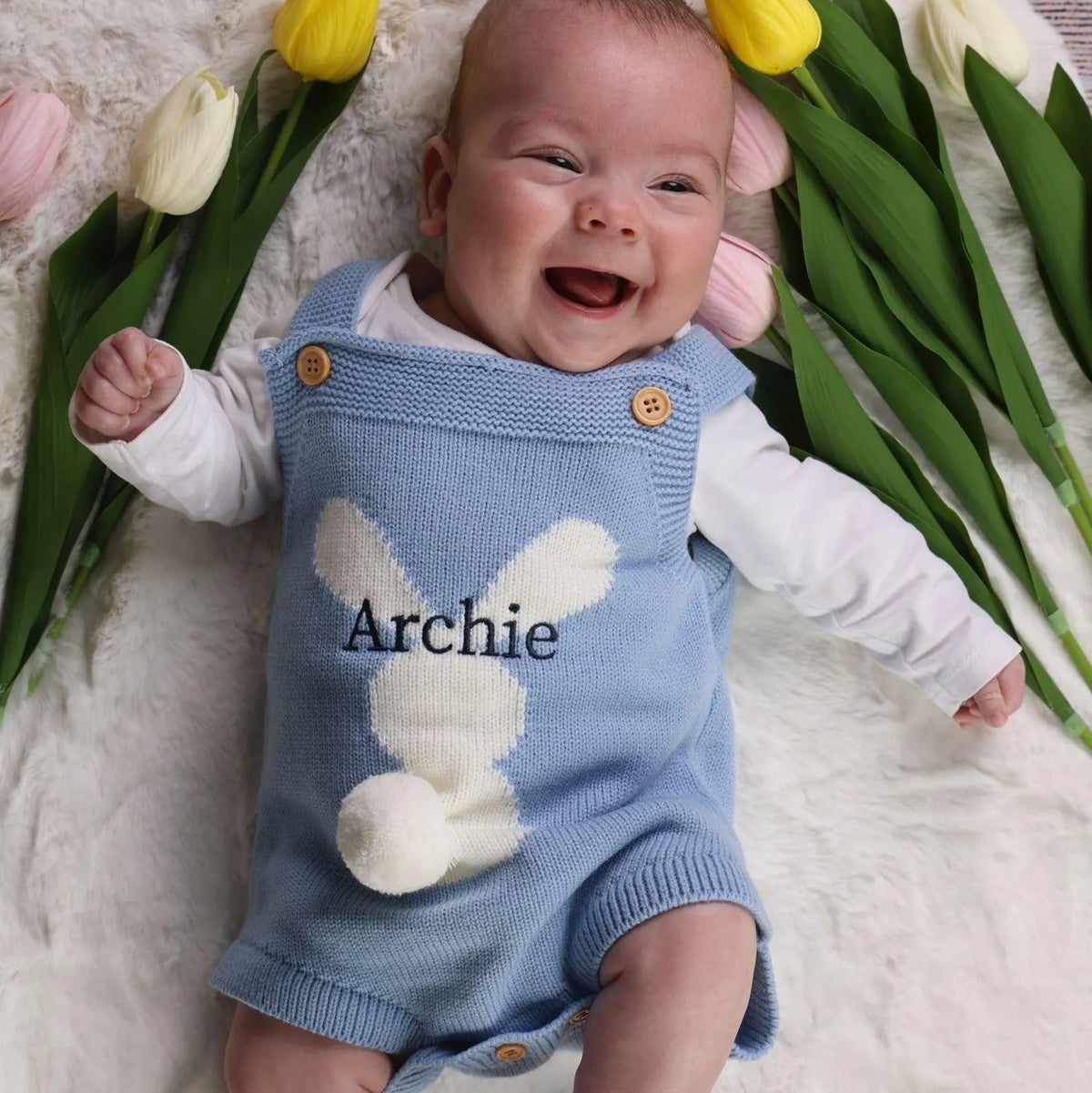 Baby in a blue knitted romper with a bunny design, surrounded by tulips on a white blanket.