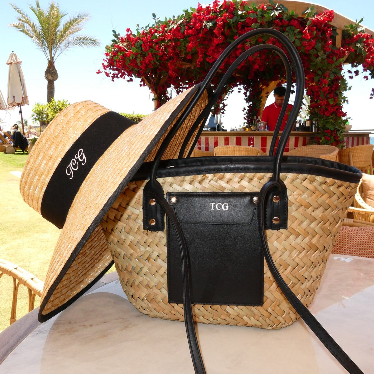 Woven straw bag with black accents and a matching hat on a beach setting.