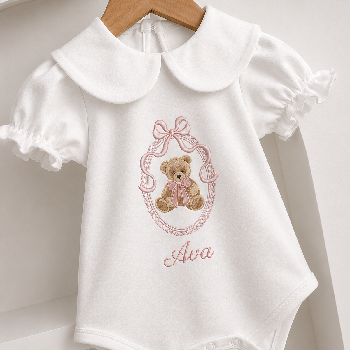 White baby bodysuit with a large Peter Pan collar, short ruffled sleeves, and an embroidered teddy bear design with the name 'Ava' in pink script, hanging on a wooden hanger.
