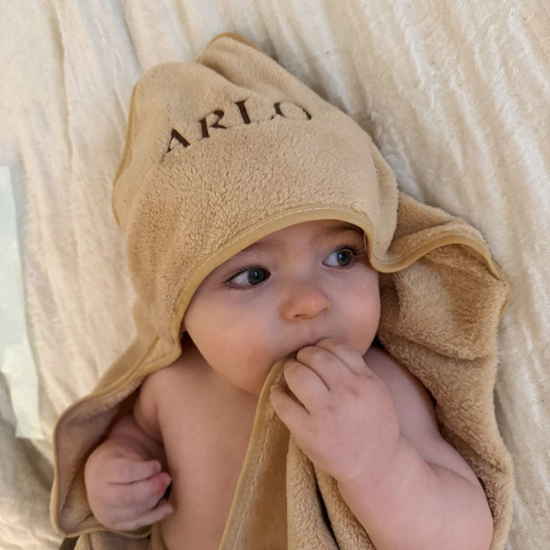 Baby wearing a beige hooded towel with 'ARLO' embroidered on it, wrapped in a soft blanket.
