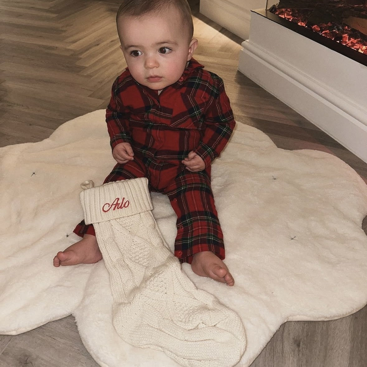 Baby in red plaid pajamas sitting on a white rug with a Christmas stocking.