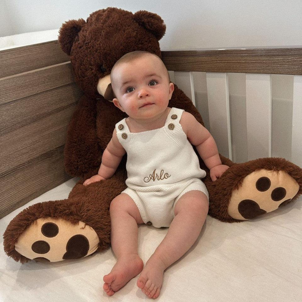 Baby in a crib with a large teddy bear, wearing a white romper with 'Arlo" embroidered.