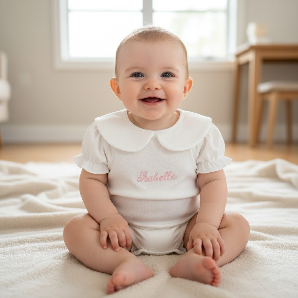 Baby sitting on a blanket wearing a white onesie with 'Isabelle' embroidered on it.